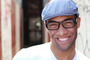A young man wearing a hat with glasses smilling with white teeth.